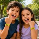 Two smiling children outdoors wear yellow Brazil wristbands and gesture excitedly toward the camera.