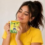 Woman in a yellow shirt smiling with eyes closed, holding a colorful mug close to her face.