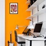 Bright orange accent wall behind a white desk with a computer, tablet stand, and two silver desk lamps. A black electric guitar rests against the wall; white shelving sits to the right.
