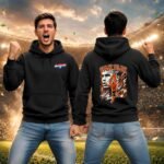 Two men in black hoodies celebrate in a stadium; one faces forward shouting while the other shows a back design reading 'VAN DIJK' with a figure and flame graphics.