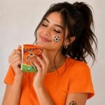 Smiling woman in an orange shirt holds a soccer-themed mug, with a small soccer ball sticker on her cheek.